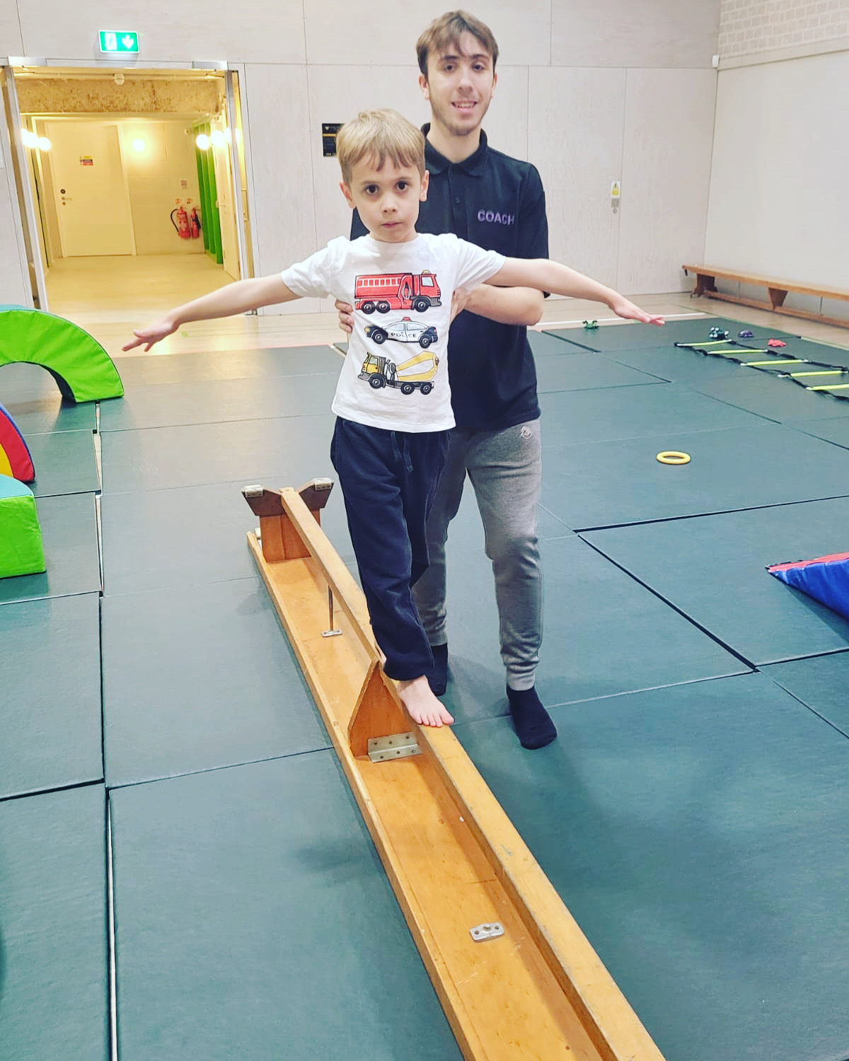 A young child balancing on a bench in the hall, with a male coach supporting him.