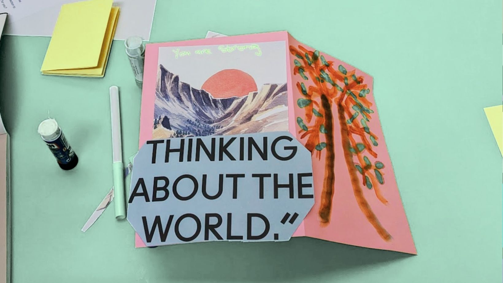 A table with art materials and a home-made zine decorated with cut-out text and imagery from a magazine reading 'THINKING ABOUT THE WORLD'. 