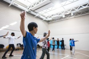 A group of adults of different ethnicities is practising Qi Gong in the large, light-filled Emerald Hall at Holborn House. 