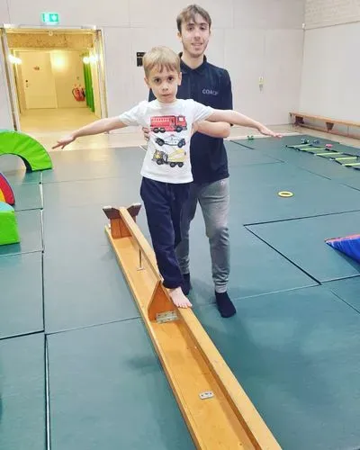 A young child balancing on a bench in the hall, with a male coach supporting him.