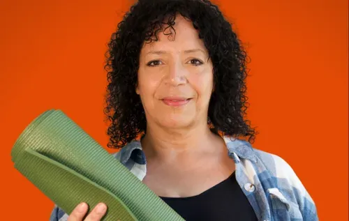 Yoga instructor, Shirley, is smiling at the camera. Shirley is a mixed-race woman with shoulder length curly, black hair. She is holding a green yoga mat and is pictured against a red background. 