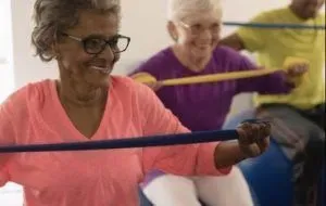 Three older adults of different ethnicities are smiling as they stretch resistance bands in front of their chests.