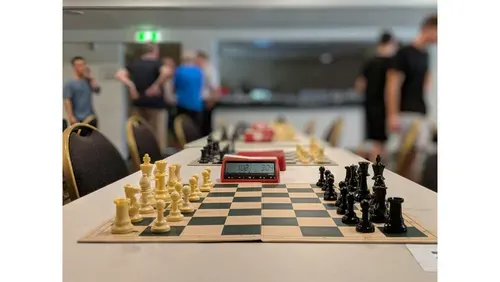 a closeup of a chess board with a red clock by it's side, in the background, other boards, and people socialising