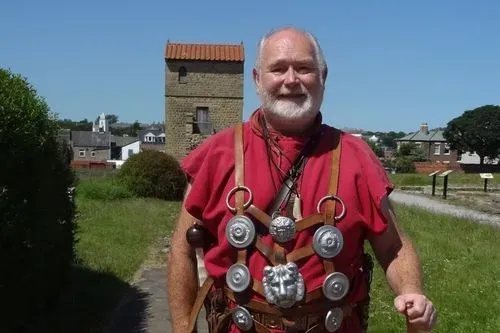 Man in a red tunic on a summers day with the reconstructed West Gate in the background