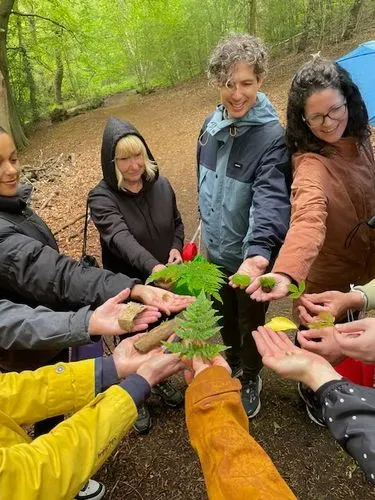 A group of people all with an arm out stretched to the centre of a circle, holding an item found in nature, within the woods.  