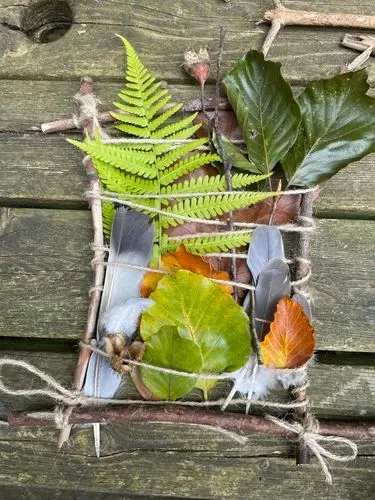 A nature weaving loom made from string and twigs with leaves, feathers and ferns woven between the string. 