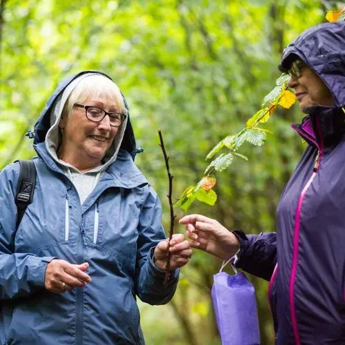 2 ladies in rain coats in the woods smiling and passing a stick between them