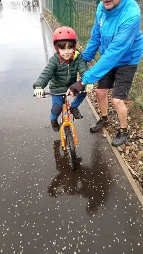 A young boy learning to ride a bike. He is smiling. An adult is helping him