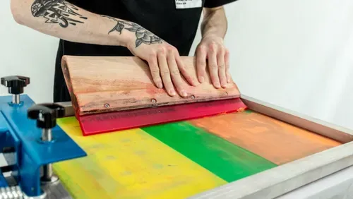 Person using a large squeegee on a colourful fabric surface including orange green yellow red. The person has tattoos on their arm and a black T-shirt on.