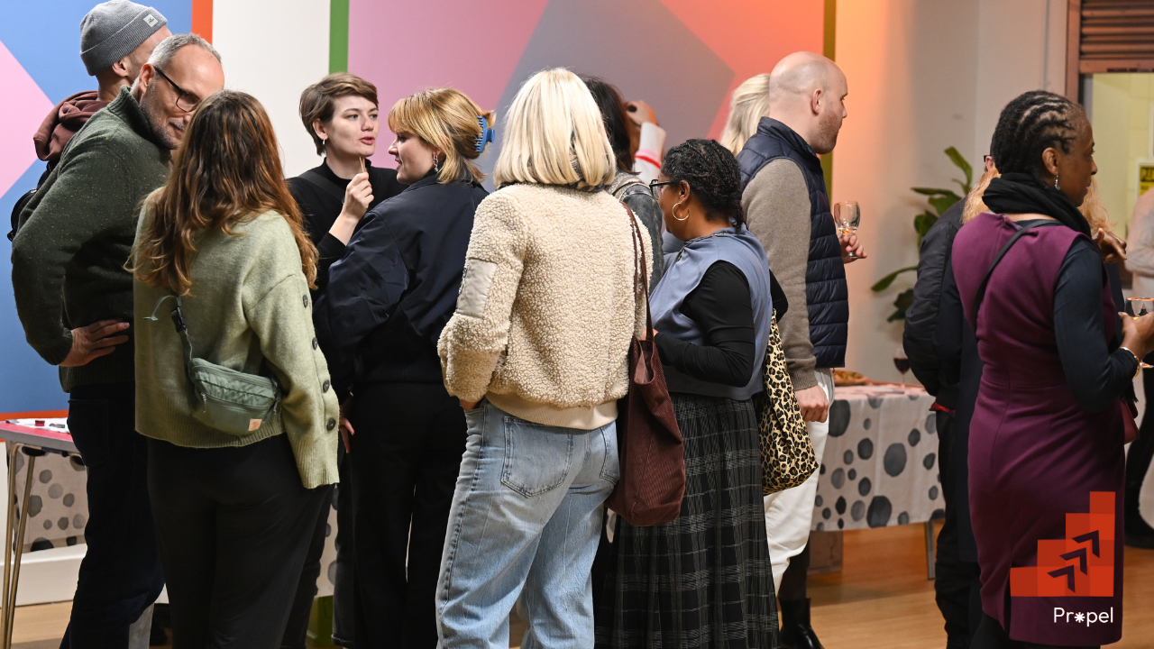 A group of people stand in small clusters, talking and networking at an indoor event. The space has colourful geometric wall art in the background and a table with food and drinks. Most attendees are holding glasses and wearing casual winter clothing. A red logo in the bottom right corner reads "Propel" with three upward arrows.