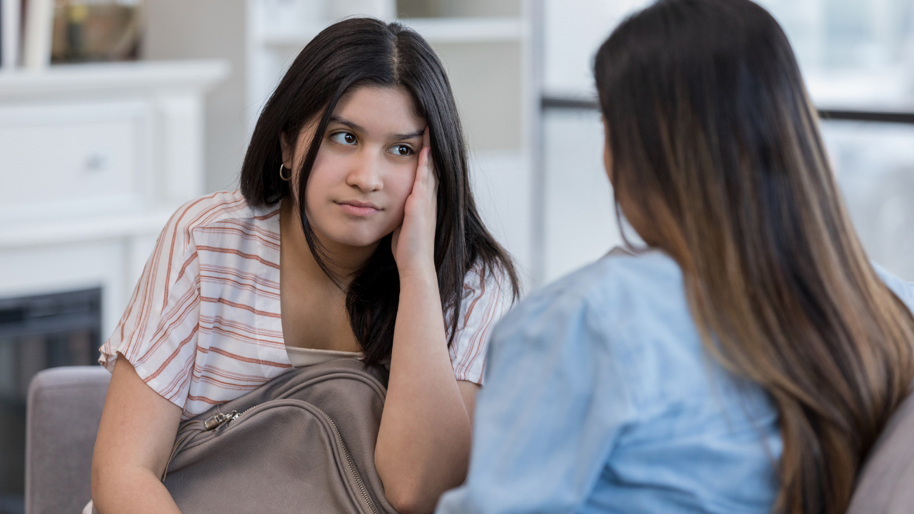 A teenage girl wearing a white and red stripy t-shirt speaks to an adult. They are deep in conversation. The adult's back is to us - she is wearing a light blue shirt and has long dark hair.