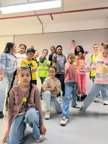A group of children in colourful clothing and adults in high visibility vests pointing to the camera / you.
