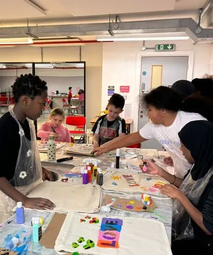 Children leaning and painting on a canvas covered table, scattered with paints pots and canvas bags.