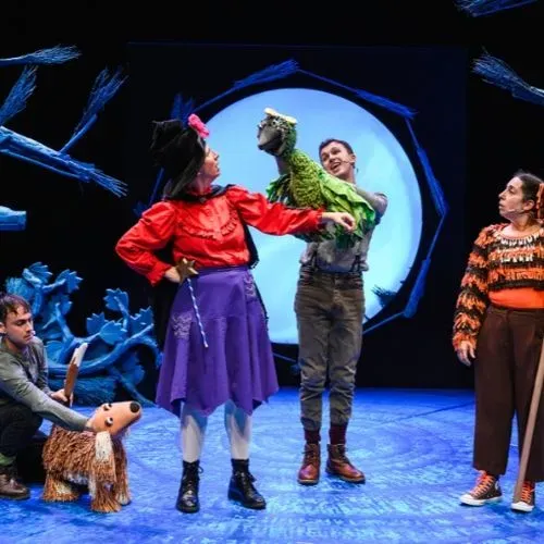 Four performers on stage one crouched down operating the Dog puppet character, a performer playing the Witch standing and performer holding the Bird puppet character and a performer as Cat.  They are on a dark blue set with brooms surrounding a circle screen and tree stumps.