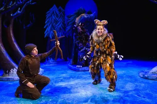 A narrator dressed in dark brown kneeling down holding stick man out to the Gruffalo's Child who is smiling.  On a set with snowy trees of the deep dark wood.