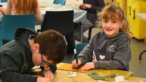 Two children are completing craft activities. One young person has their head down focused on drawing. The other child is smiling directly at the camera.