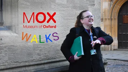 A photo of tour guide Hannah Field standing in front of a building in Oxford. She is wearing a black coat and a rainbow lanyard and holding a green folder.