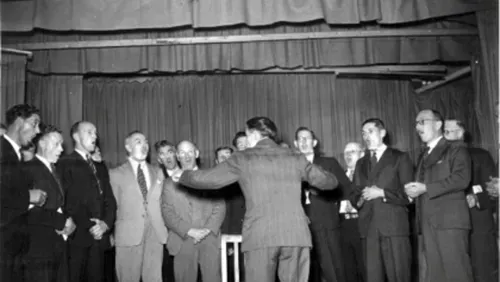 A black and white photograph of a group of men wearing suits standing in front of a stage, singing. They are being led by a conductor, who is gesturing enthusiastically in the centre.
