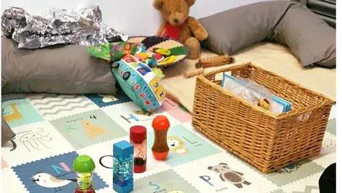 Play corner set up on a mat, with some children's toys, including; sand timers, books, a teddy, a silver foil blanket and a shaker. 