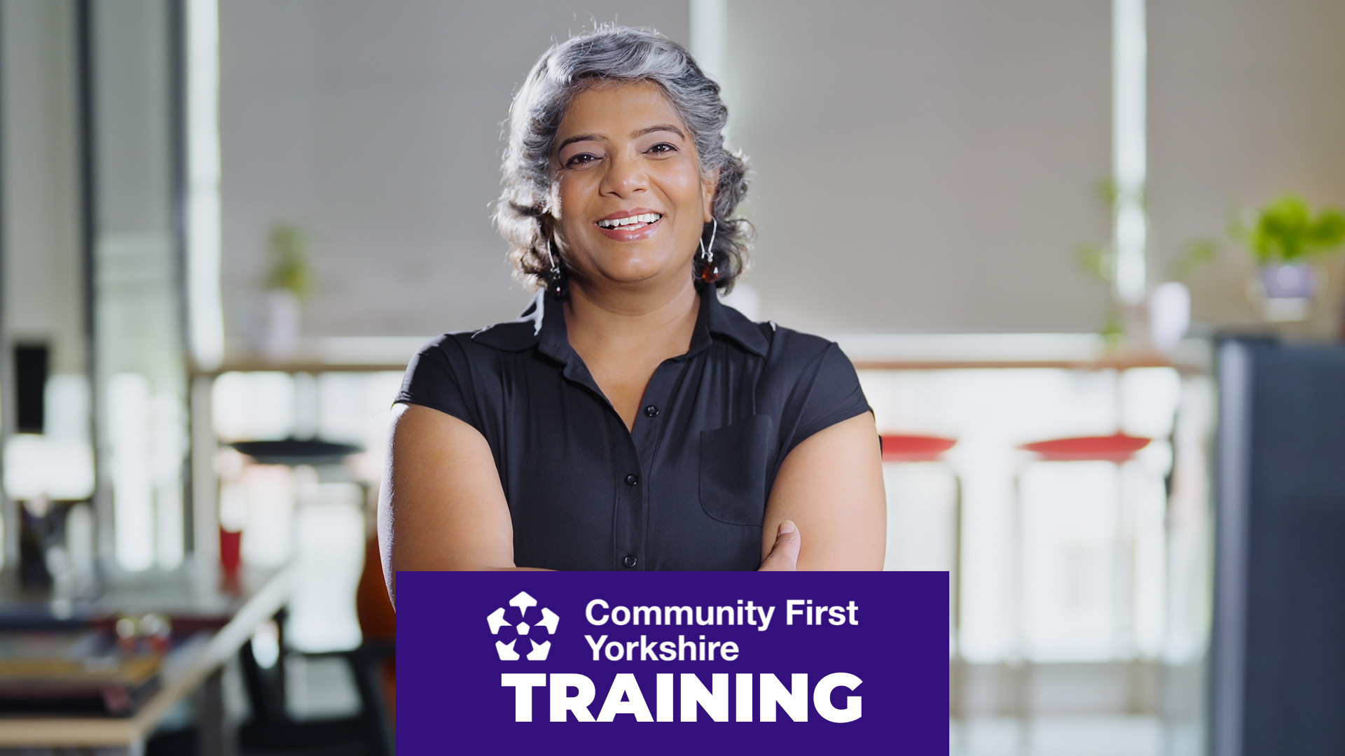 Person standing in an office environment with arms crossed, wearing a short-sleeved black shirt. Background shows desks, chairs, and large windows. Purple banner reads “Community First Yorkshire TRAINING.”