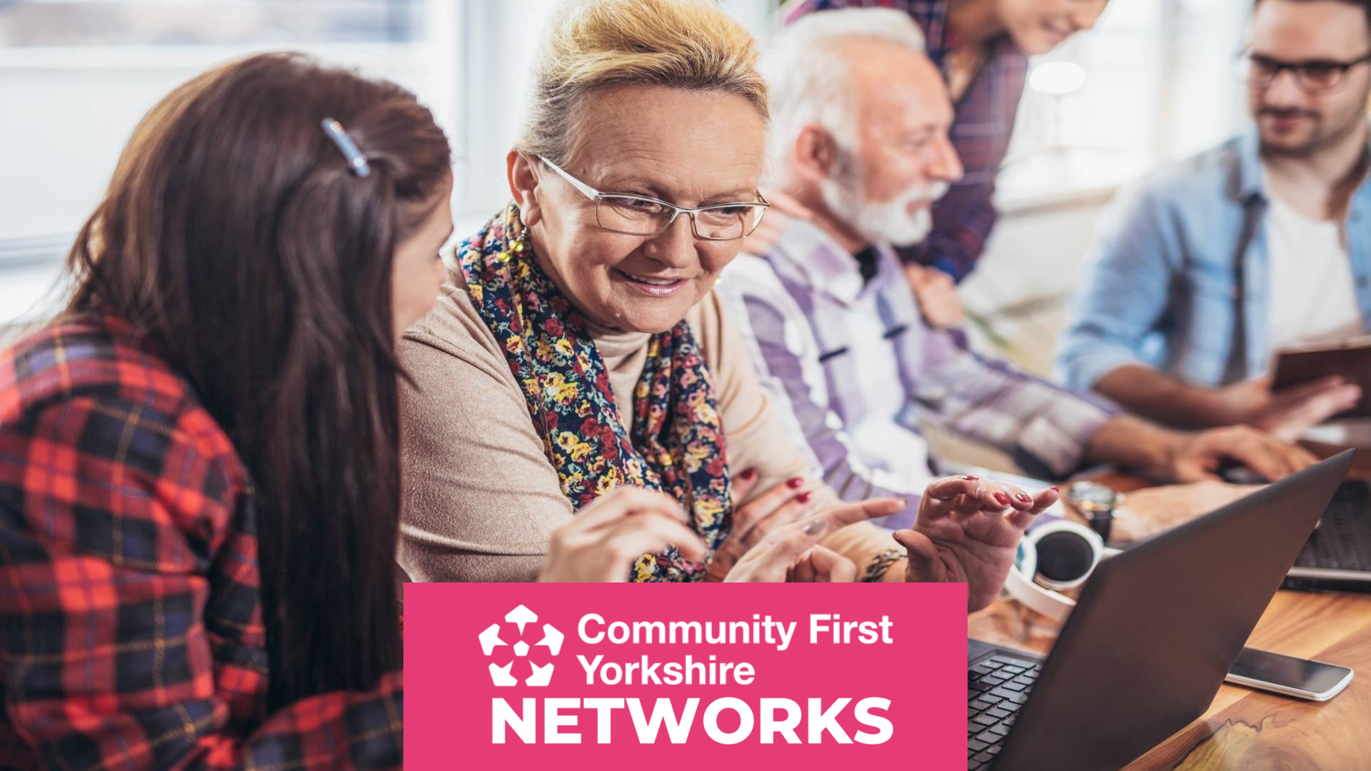 Group of people sitting around a table with laptops and notebooks, engaged in discussion. Pink banner reads “Community First Yorkshire NETWORKS.”