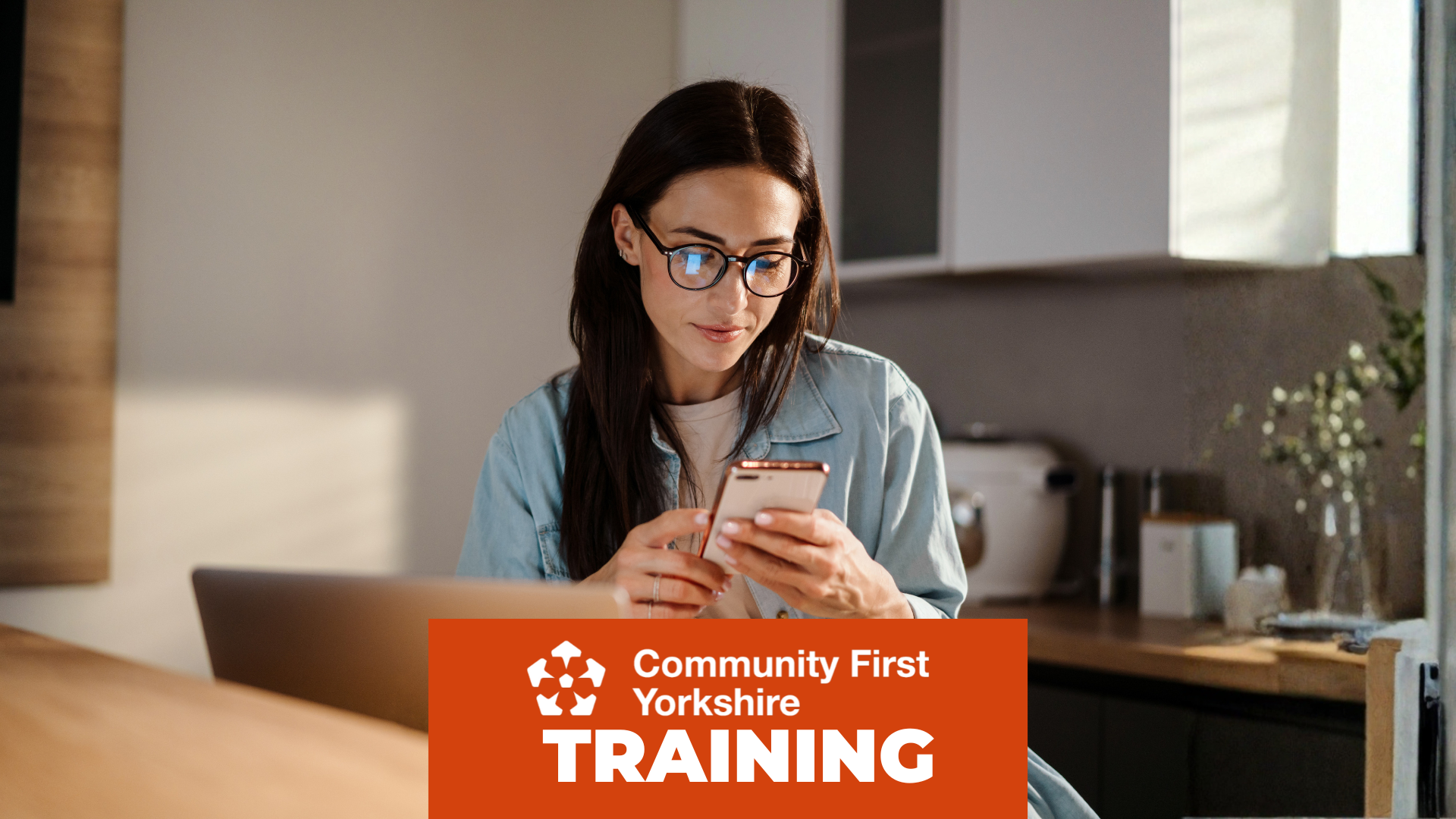 Person sitting at a table holding a smartphone, with a laptop in front. Orange banner reads “Community First Yorkshire TRAINING.”