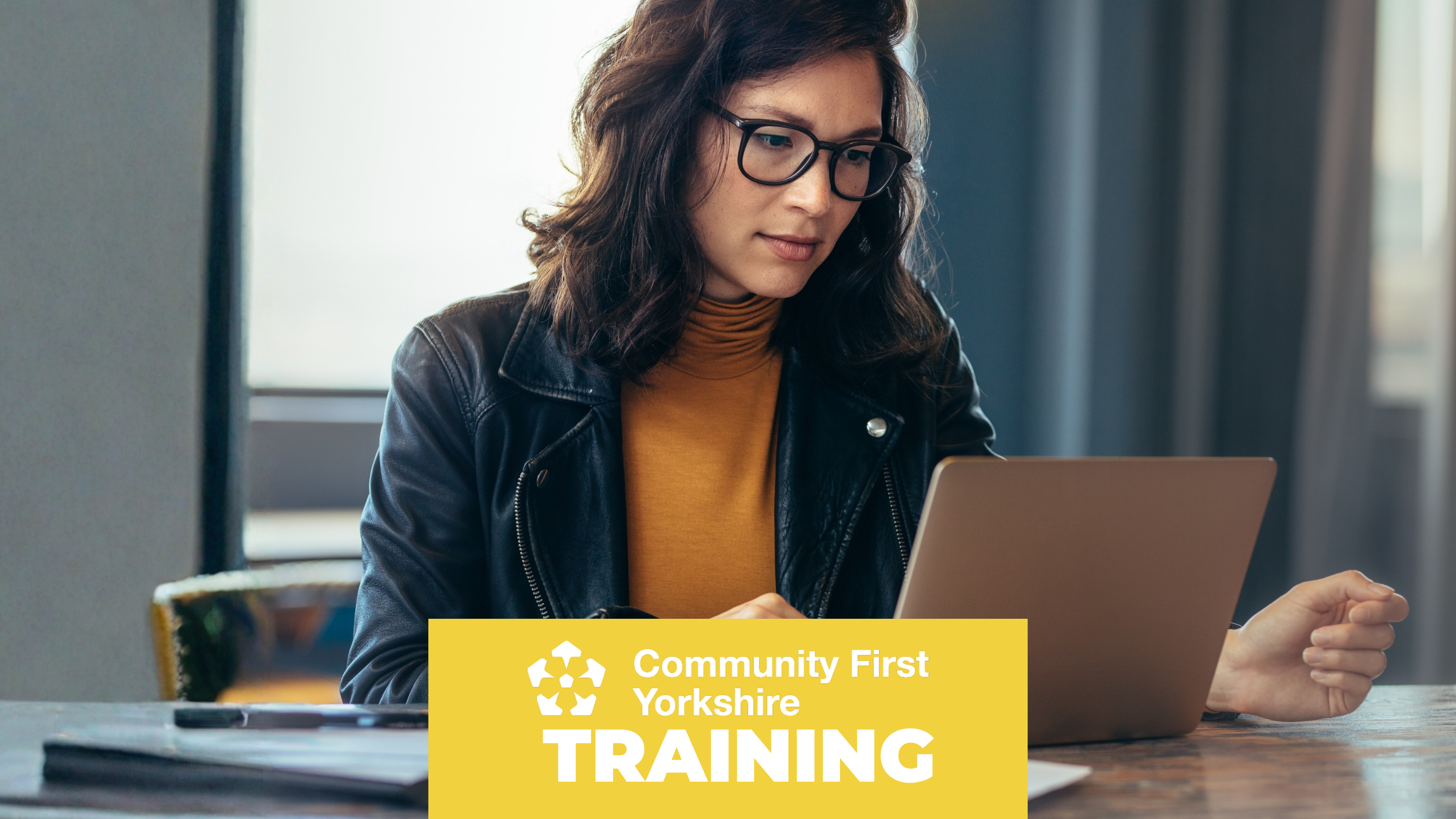 Person sitting at a table using a laptop, wearing a black jacket and an orange jumper. Yellow banner reads “Community First Yorkshire TRAINING.”