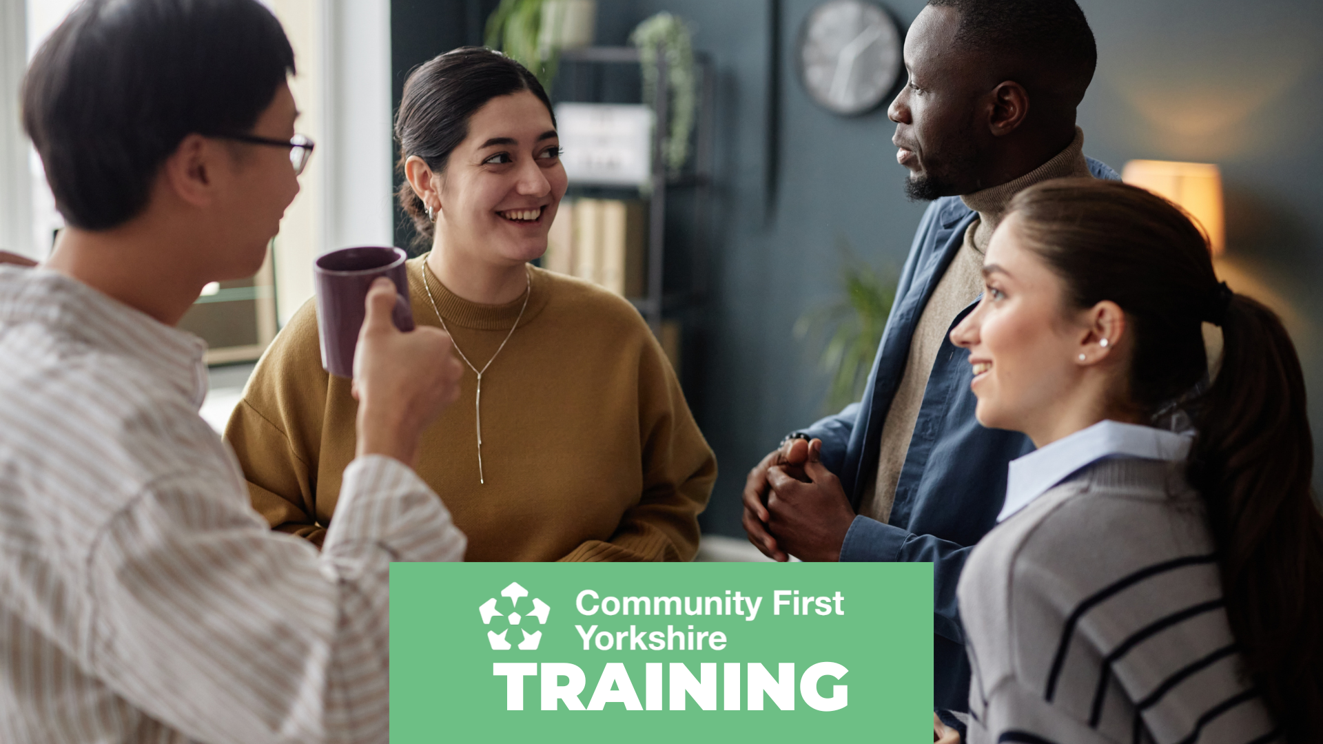 Four people standing and talking in an indoor setting with shelves and a clock in the background. One person is holding a mug. Green banner reads “Community First Yorkshire TRAINING.”