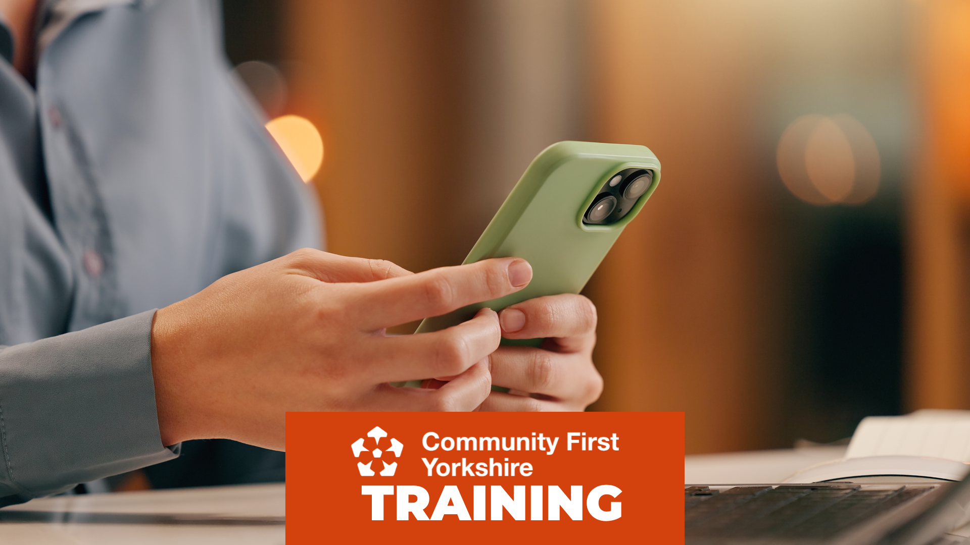 Hands holding a green smartphone above an open notebook and a laptop on a desk. Orange banner reads “Community First Yorkshire TRAINING.”