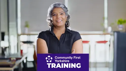 Person standing in an office environment with arms crossed, wearing a short-sleeved black shirt. Background shows desks, chairs, and large windows. Purple banner reads “Community First Yorkshire TRAINING.”