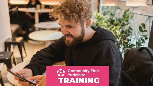 Person sitting in a café or informal workspace, looking at a smartphone beside an open laptop, with Community First Yorkshire Training branding displayed at the bottom of the image.