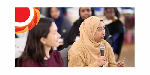 Woman in a tan hijab holds a microphone and speaks during a group discussion, while other participants sit blurred in the background. THCVS logo in the top left corner.
