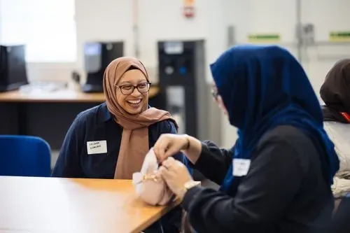 In this image there is a woman in a beige hijab and glasses looking over at another hijabi woman who is also laughing