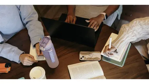 Overhead view of a study or work session at a wooden table, with a laptop in the center, two people's hands visible, a water bottle, a coffee cup, an open book, notebooks, a phone, and a pen.