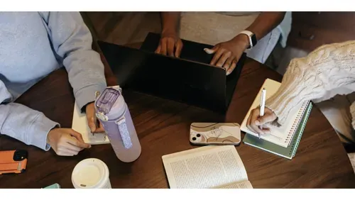 Overhead view of three people gathered around a wooden table, working together. One person types on a black laptop in the centre, another writes in a notebook while holding a water bottle, and a third writes in a spiral notebook with a pen. A smartphone with a line-drawing case sits on the table alongside the notebooks.