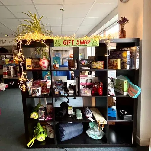 Inside of a shop, there is shelve unit full of potential presents. There is a decorative plan on top anda label reading GIFT SWAP