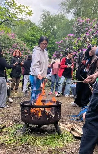 A young person is toasting a marshmallow on a campfire. Other young people and youth workers are gathered round